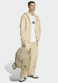Beige tracksuit with hooded jacket and matching pants, white t-shirt with black logo, carrying a beige textured backpack with multiple pockets.