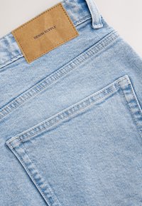 Jean en denim bleu clair avec une étiquette en cuir portant l'inscription "DENIM SUPPLY", doté de poches droites et de détails de couture visibles.