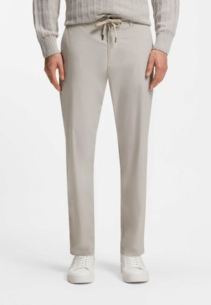 Mężczyzna w jasnobeżowych spodniach z wiązaniem, beżowym prążkowanym swetrze i białych trampkach, stojący na tle gładkiej białej ściany.