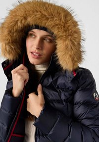 Giaccone imbottito blu navy con ampia cappuccio in pelliccia sintetica marrone chiaro, con chiusura a zip e dettagli in rosso, indossato sopra un top color crema.