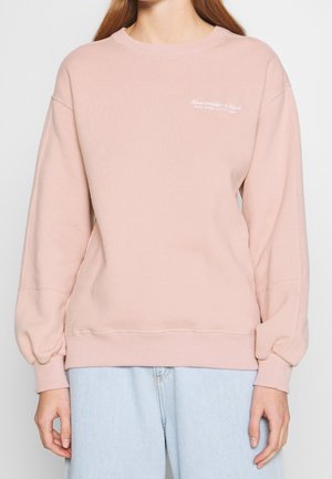 Person trägt ein hellrosa Abercrombie & Fitch Sweatshirt mit kleinem weißem Logo auf der Brust und hellblaue Jeans.