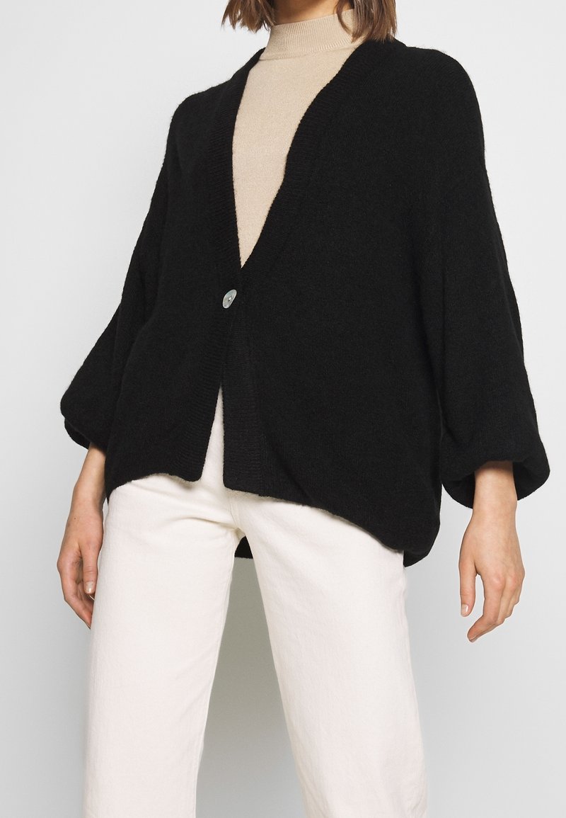 Schwarzer oversized Cardigan aus weichem Material mit tiefem V-Ausschnitt, einreihiger Knopfleiste und weiten Ärmeln. Getragen über einem beigen Oberteil.