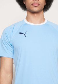 Camiseta atlética de color azul claro, fabricada en un material suave y transpirable. Presenta un cuello redondo blanco y un logo de Puma en azul oscuro en la parte superior izquierda.