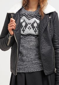 Donna che indossa una giacca di pelle trapuntata nera con colletto in pile bianco sopra un maglione a maglia con motivo in bianco e nero, mentre tiene il risvolto della giacca.