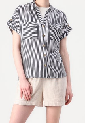 Person wearing short-sleeve black and white vertical striped button-up shirt over a light-colored top, paired with loose beige shorts.