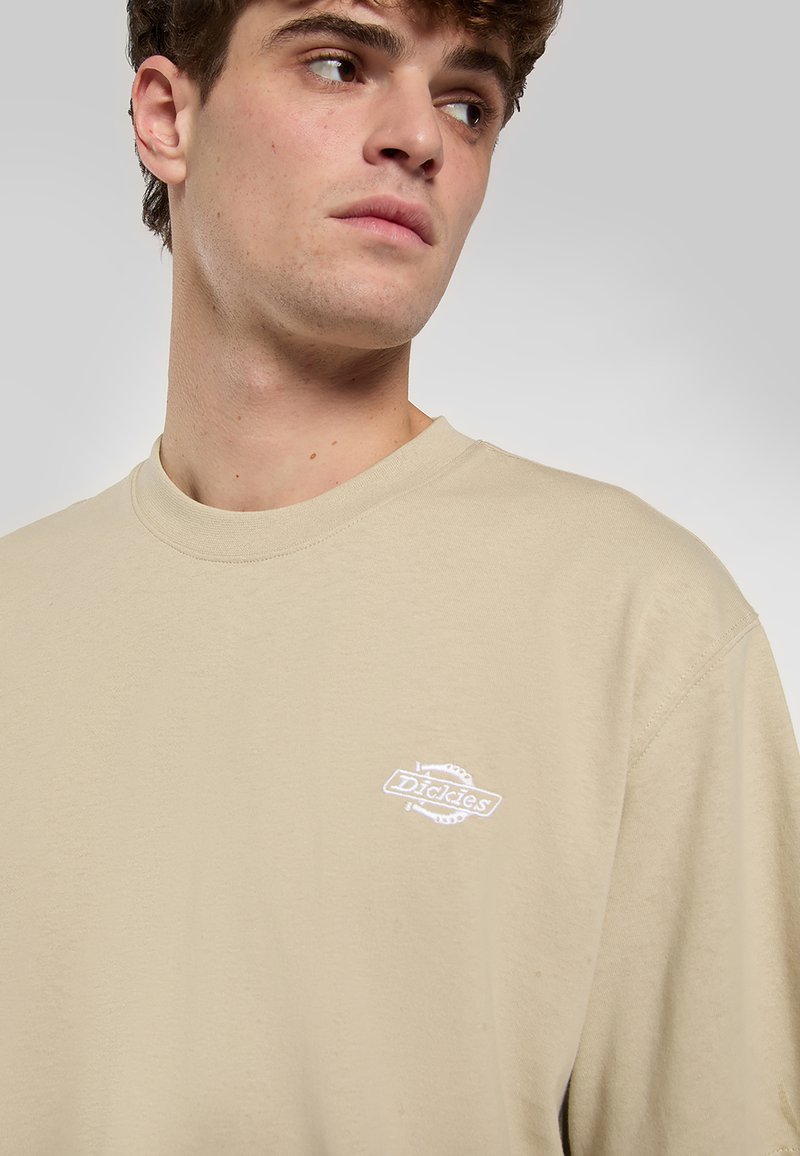 Beige katoenen T-shirt met een ronde hals en een klein wit geborduurd logo op de linkerborst, met een ontspannen pasvorm en een gladde textuur.