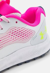 Chaussure de sport blanche avec des accents roses et une tige en mesh, dotée d'une semelle texturée et d'un design dynamique. Détail de logo réfléchissant sur le côté.