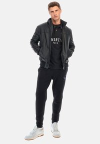 Svart läderbombarjacka över en svart pikétröja med logotyp, kombinerad med svarta sweatpants och vita sneakers. Skräddarsydd passform, avslappnad design.