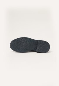 Suela de zapato de goma negra con una superficie texturizada y punta redondeada, que presenta un pequeño acento rectangular cerca del talón.