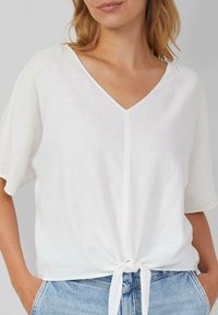 Weißes Leinenoberteil mit V-Ausschnitt, kurzen Ärmeln und einer vorderen Bindedetail. Weiche Textur und lockere Passform, kombiniert mit Jeans.