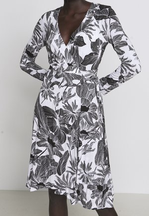 Mujer con un vestido cruzado floral blanco y negro, de manga larga y escote en V, de pie con las manos en las caderas sobre un fondo liso.