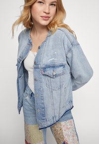 Lichtblauwe spijkerjacket met een ontspannen pasvorm, voorzien van twee borstzakken, knoopsluiting en vervaagde details. Draagt over een wit tanktop.
