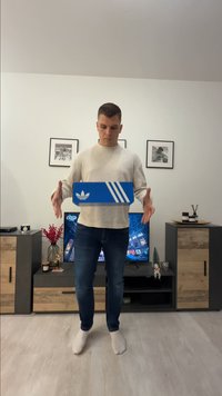 Mies valkoisessa villapaidassa ja farkuissa leijuttaa sinistä Adidas-kengännysrasiota olohuoneessa, jossa on televisio, kaapit ja seinällä kehystettyjä valokuvia.