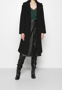 Manteau noir texturé avec un large col, associé à un haut en satin vert et un pantalon en cuir noir. Les bottes sont également noires et ajustées.