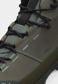 Κοντινό πλάνο πεζοπορικού παπουτσιού ελιάς πράσινου χρώματος με μαύρα κορδόνια, επενδυμένη γλώσσα, ορατές ραφές και τη σήμανση «POWERTEK» στο πλάι.