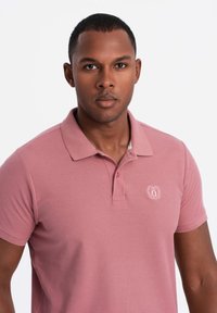 Vaaleanpunainen lyhythihainen polo-paita, jossa on kaulus, pieni kirjailtu logo rinnassa ja sileä, teksturoitu kangas.