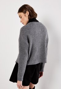 Frau mit braunem, kurzen Haaren, die einen gerippten grauen Pullover über einem schwarzen Kleid trägt und seitlich an einer schlichten weißen Wand steht.