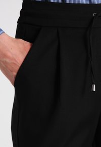 Pantaloni neri con vita elastica e laccetto, con pieghe e tasche laterali. Materiale liscio con finitura opaca. Camicia a righe visibile sopra.