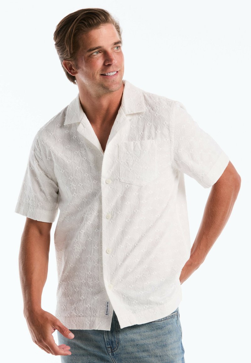 Man smiling, wearing a short-sleeve white textured button-up shirt and blue jeans, with one hand on hip and looking to side.