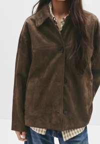 Hnědá semišová bunda s špičatým límcem, zapínáním na dva knoflíky a boční kapsou; nošena přes kostkovanou košili a džíny.