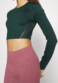 Vihreä pitkähihainen crop-top, jossa on mustat korostusraidat sivuilla, yhdistettynä korkeavyötäröisiin vaaleanpunaisiin leggings-housuihin, joissa on pieni logo.