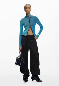 Cardigan a costine con zip in blu brillante abbinato a pantaloni cargo neri ampi. Il modello tiene una piccola borsa nera e indossa stivali a punta.