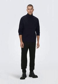 Pulôver de gola alta azul-marinho com textura canelada, combinado com calças pretas e botas pretas robustas. O modelo está posicionado contra um fundo liso.