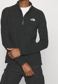 Svart fleece-pullöver med zipkrage, prydd med en vit logotyp på vänster bröst och tonande sömmar längs ärmarna. Tyget verkar vara mjukt.