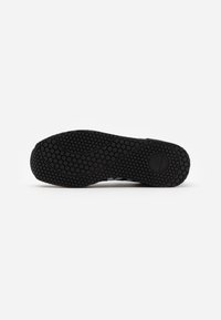 Svart såle på sneakers med et teksturert heksagonalt mønster og buede linjer, med en rund dekorasjon nær hælen.