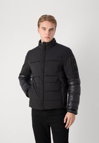 Black puffer jacket with a high collar, contrasting matte and shiny textures, quilted design, zippered side pockets, and a sleeve pocket.