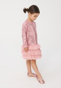 Abito rosa con bustino di paillettes e volant di tulle a strati, abbinato a ballerine rosa con un accento a fiocco. Bambina in piedi di profilo.