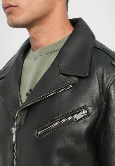 Μαύρο δερμάτινο μπουφάν μοτοσικλέτας με ασημένιες λεπτομέρειες, εσοχή στον γιακά, διαγώνιες τσέπες με φερμουάρ και υφή. Φορεμένο πάνω από ένα πράσινο φούτερ.