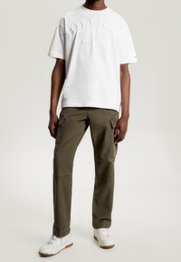 Camiseta blanca de algodón con logotipo en relieve, combinada con pantalones cargo verdes olivo con bolsillos laterales y zapatillas blancas con suela de goma.
