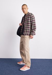 Giacca a quadri in tonalità viola e verde sopra una maglietta nera, abbinata a pantaloni beige e scarpe da ginnastica arancioni con strisce bianche. Borsa nera tenuta in mano.