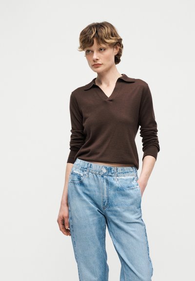 Γυναίκα με κοντά κυματιστά μαλλιά που φορά καφέ μακρυμάνικο πουκάμισο με γιακά και ανοιχτόχρωμο τζιν ψηλόμεσο παντελόνι, στέκεται με το ένα χέρι στην τσέπη.