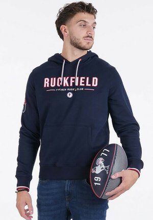 Hoodie bleu marine avec poche frontale, arborant le texte "RUCKFIELD" en blanc et rouge. Poignets et ourlet côtelés, porté avec un jean en denim, tenant un ballon de rugby.