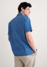 Camicetta azzurra a maniche corte con motivo a griglia e punti verdi. Realizzata in tessuto leggero, presenta un colletto casual e orlo dritto.