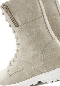 Botín de tela beige que presenta una superficie texturizada, cordones en la parte delantera con ojetes plateados y una cremallera lateral para un fácil acceso. Suela blanca.