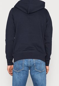 Felpa navy con cappuccio e cordino, tessuto morbido e tasca a marsupio, indossata con jeans in denim blu dotati di due tasche posteriori.