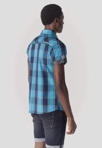 Mężczyzna w niebieskiej i granatowej koszuli w kratę z krótkim rękawem oraz ciemnych dżinsowych krótkich spodenkach podwiniętych na dole, stojący bokiem na jednolitym tle.