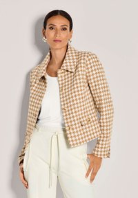Vrouw in een beige en witte houndstooth jas, witte top, crèmekleurige broek met drawstring en decoratieve oorbellen tegen een effen achtergrond.