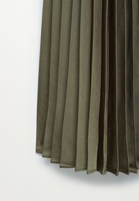 Jupe plissée vert olive suspendue sur un fond blanc, montrant une texture de tissu lisse et des plis verticaux pliés.