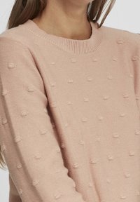 Pull léger rose avec un col rond et un motif texturé en points surélevés. Tissu en maille douce avec des poignets et un ourlet côtelés.