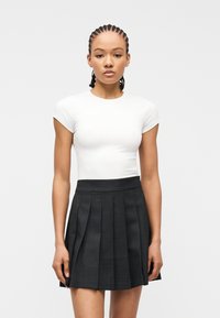 Νεαρή γυναίκα με πλεξούδες φοράει εφαρμοστό λευκό τοπ με κοντά μανίκια και μαύρη πλισέ φούστα ψηλόμεση, στεκόμενη μπροστά σε απλό φόντο.