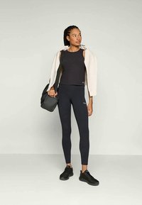 Top sem mangas atlético preto e leggins, combinados com uma jaqueta de cor clara. O conjunto apresenta um design elegante e um tecido texturizado. Ténis pretos e uma mala.
