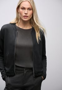 Černá bomber bunda se zipem, žebrovanými manžetami a bočními kapsami, nošená přes tmavě šedé tričko a antracitové kalhoty.