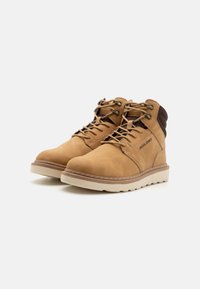 Botas de cuero marrón claro con textura suave, cordones marrones y cuello acolchado. Cuenta con una suela de goma blanca y detalles de costura en los laterales.