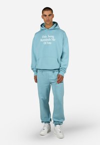 Felpa leggera blu con testo bianco, tasca a marsupio frontale e polsini a costine; pantaloni coordinati con vita elasticizzata; indossati con sneakers bianche.