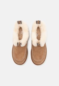 Pantuflas de ante marrón con un forro esponjoso de color crema y acentos decorativos de patrón en los puños. Detalles cosidos a lo largo de los bordes.