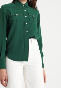 Camicia verde con colletto, due tasche sul petto, dettagli in cucitura bianca, maniche lunghe e texture liscia, abbinata a pantaloni bianchi.
